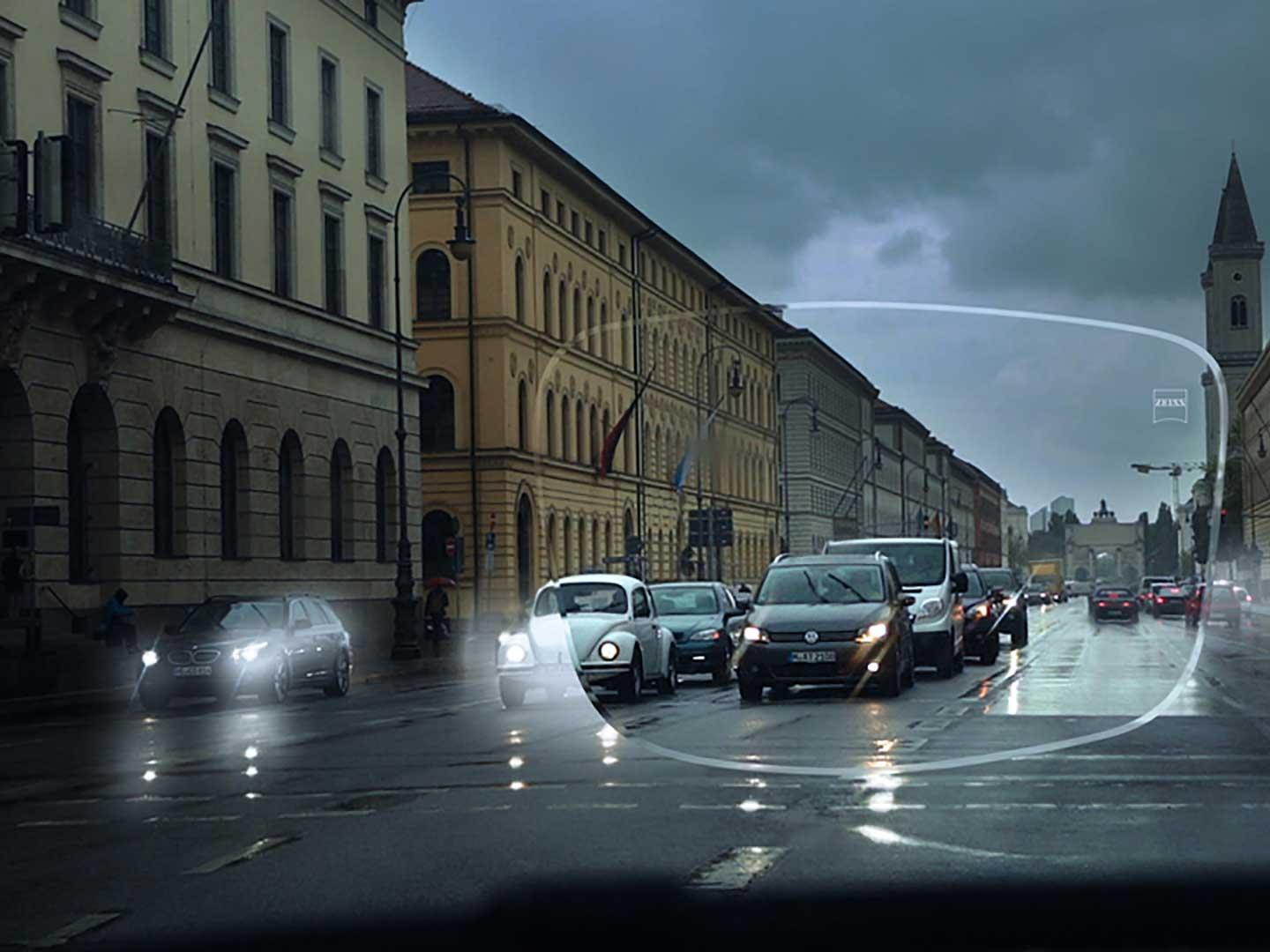 Görüntü, bir caddede düşük ışık koşullarında zayıf görüşü gösterir. Bakış açısı, bir gözlük camından görülen bir arabanın içidir. 
