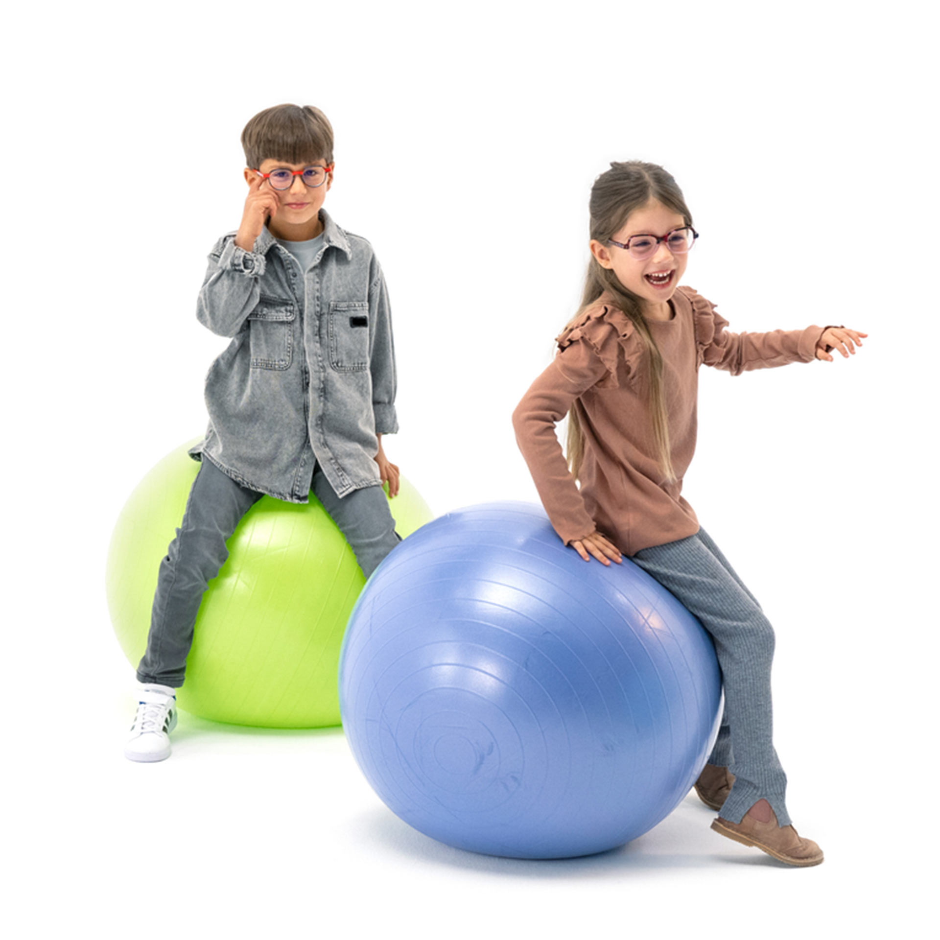 Her ikisi de gözlük takan bir erkek ve bir kız çocuk, spor toplarının üzerinde eğlenerek zıplıyorlar.