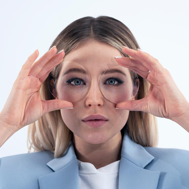 Genç sarışın bir kadın, ZEISS ClearView tek odaklı camlar nedeniyle gözlerinde herhangi bir komik etki yaratmadan iyi görünüm sergilemek için gözlerinin önünde cam tutuyor.