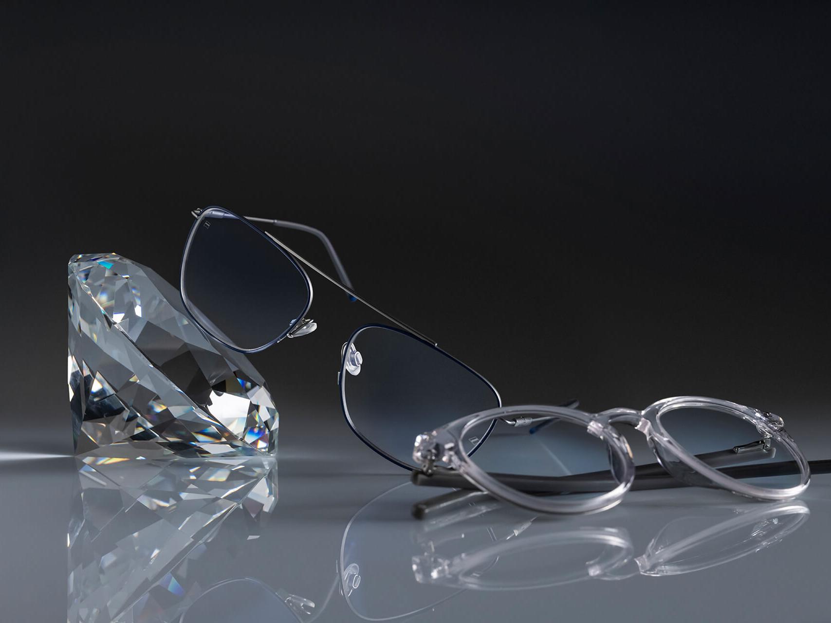Şimdiye kadarki en sert ZEISS kaplaması olan DuraVision® Platinum'a sahip ZEISS camlı iki gözlük. Bir bardak kristale yaslanmış, diğeri ise yerde düz bir şekilde duruyor. Her ikisi de mavi yansıma yapmayan şeffaf camlara sahiptir.