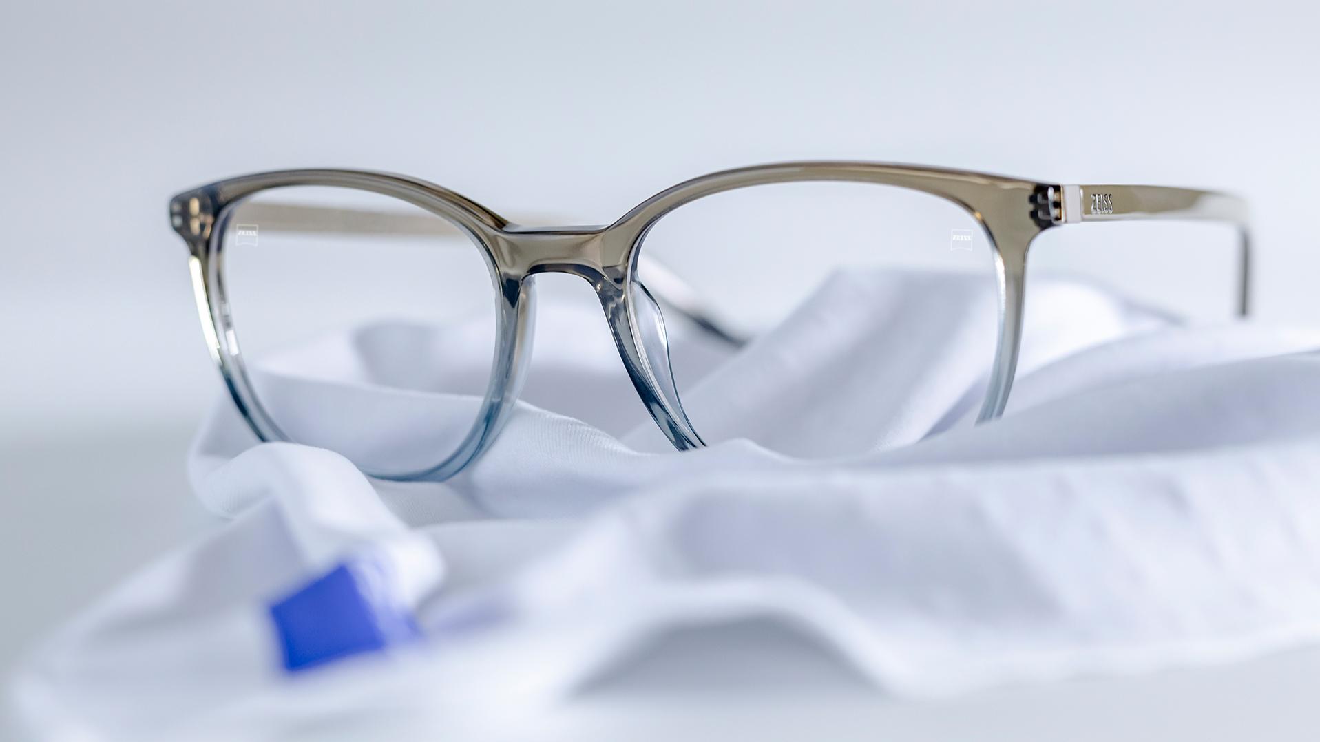 Gri-mavi çerçeveli ve DuraVision® kaplamalı ZEISS camlı bir gözlük, beyaz bir mikrofiber bezin üzerinde duruyor.
