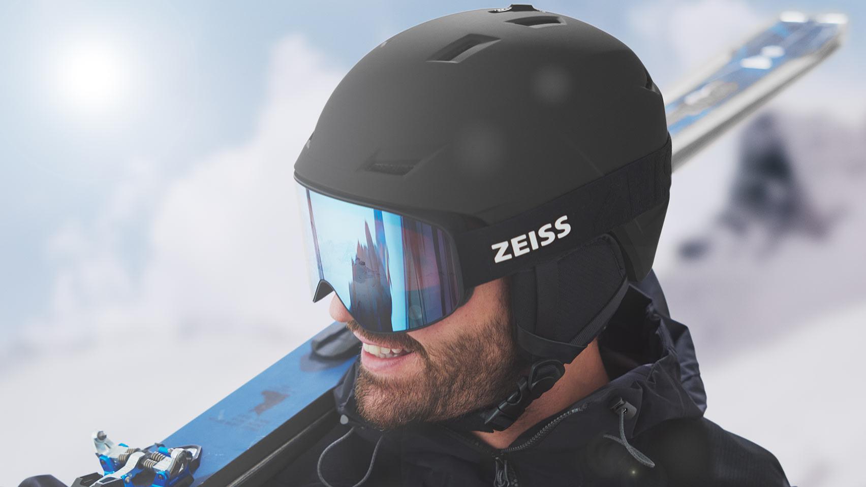 Bir adam siyah bir kask ve mavi aynalı ZEISS kayak gözlüğü takıyor, kayakları sağ omzunda taşıyor.